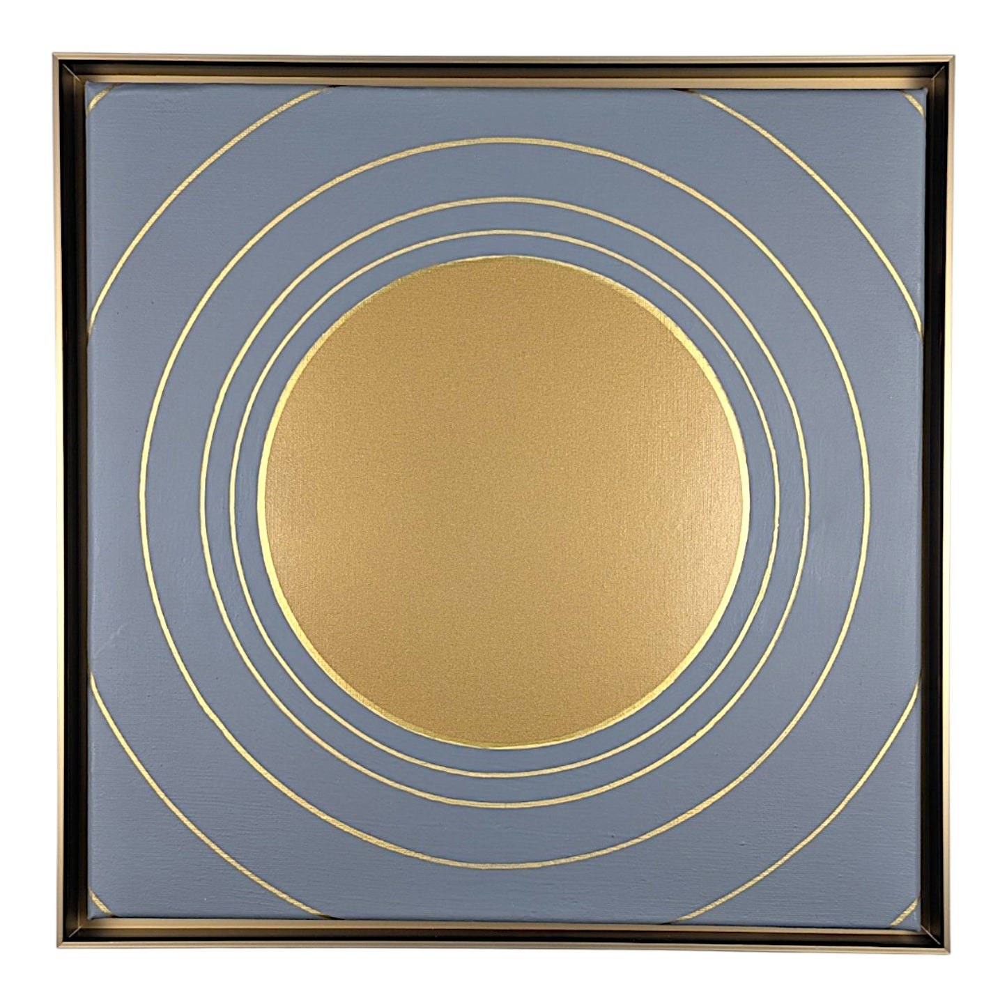 Square framed wall art featuring a large metallic gold circle at the center with multiple thin gold concentric rings on a muted blue-gray background, surrounded by a dark frame.