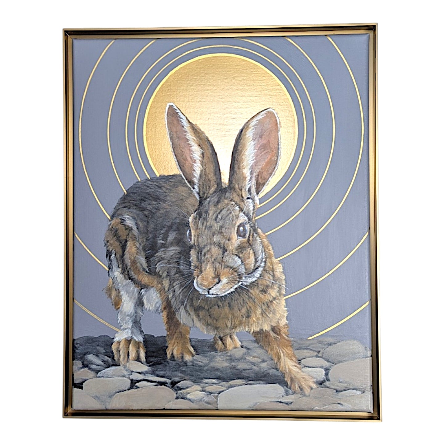 A framed painting of a brown and gray rabbit standing on a rocky surface, with a large gold circular sun and concentric gold rings on a muted blue-gray background. The artwork is enclosed in a thin gold frame and shown against a plain white backdrop.